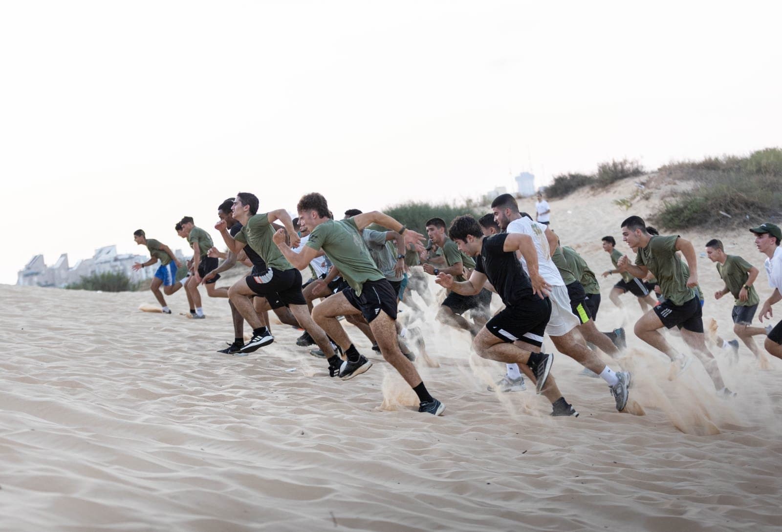 קבוצה גדולה של חניכי אדרנלין מזנקת בריצה במעלה דיונה, וחול עף מתחת לרגליהם