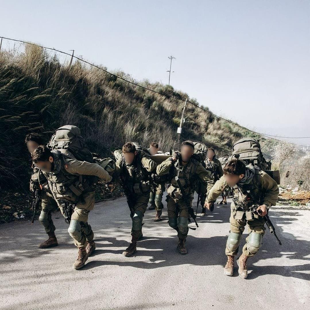 לוחמי אגוז בציוד מלא ובתיקי גב נושאים אלונקה בריצה על כביש עולה, ופניהם מטושטשים