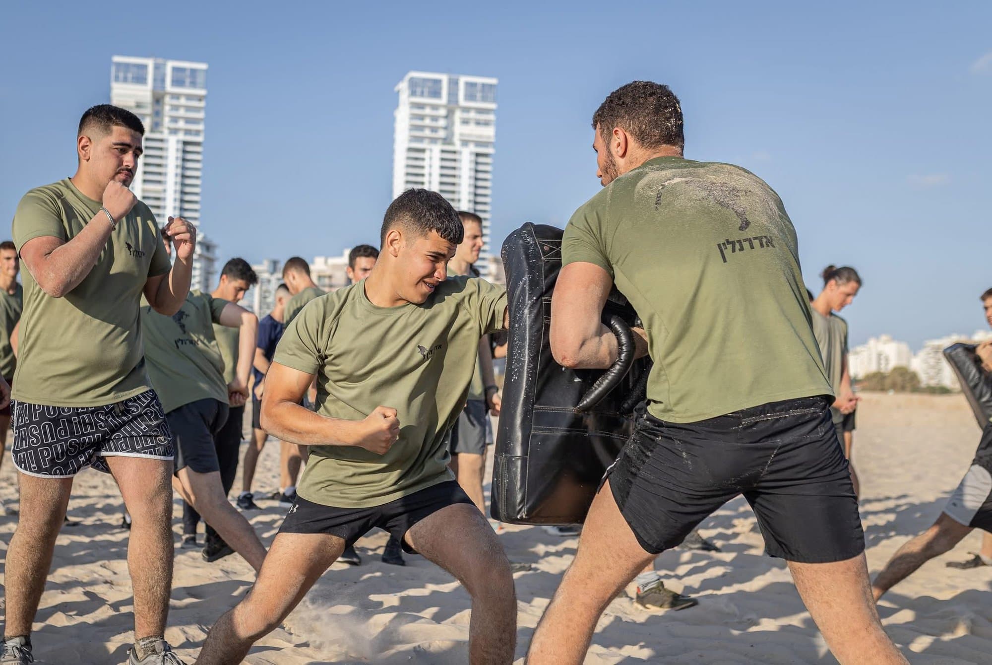 חניך בחולצה ירוקה של אדרנלין כושר קרבי מתרגל מכת אגרוף בכרית אימון שמחזיק אדם אחר, על חוף הים, וחניכים נוספים עומדים סביבם