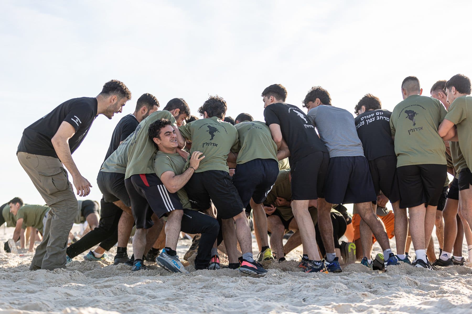 חניכי אדרנלין כושר קרבי נדחקים יחד בגוש צפוף על החול, ומדריך עומד לצידם