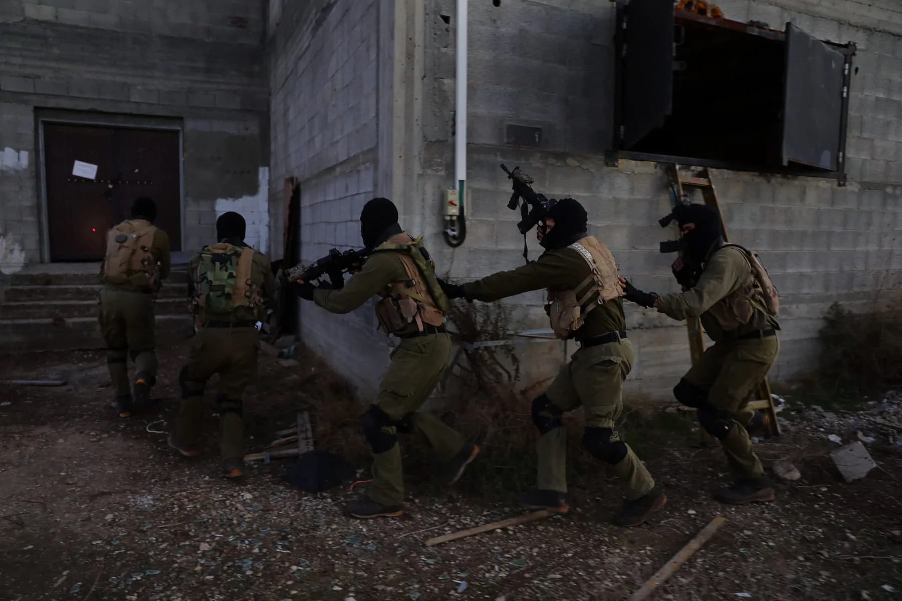 צוות לוחמים רעולי פנים מתקדם בטור לאורך קיר של מבנה, בתרגיל לחימה בשטח בנוי
