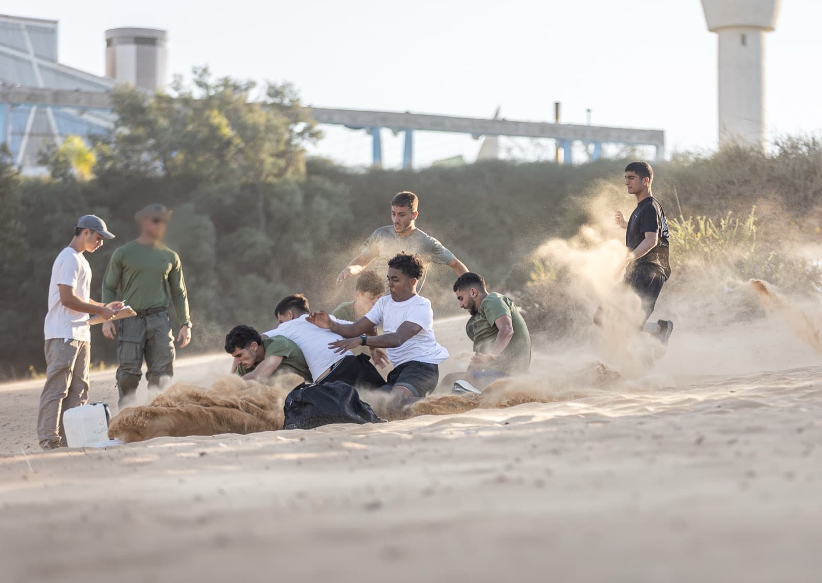 חניכי אדרנלין כושר קרבי מזנקים אל שקי חול בתוך ענן אבק, ולצידם עומדים מדריך ואדם שרושם תוצאות