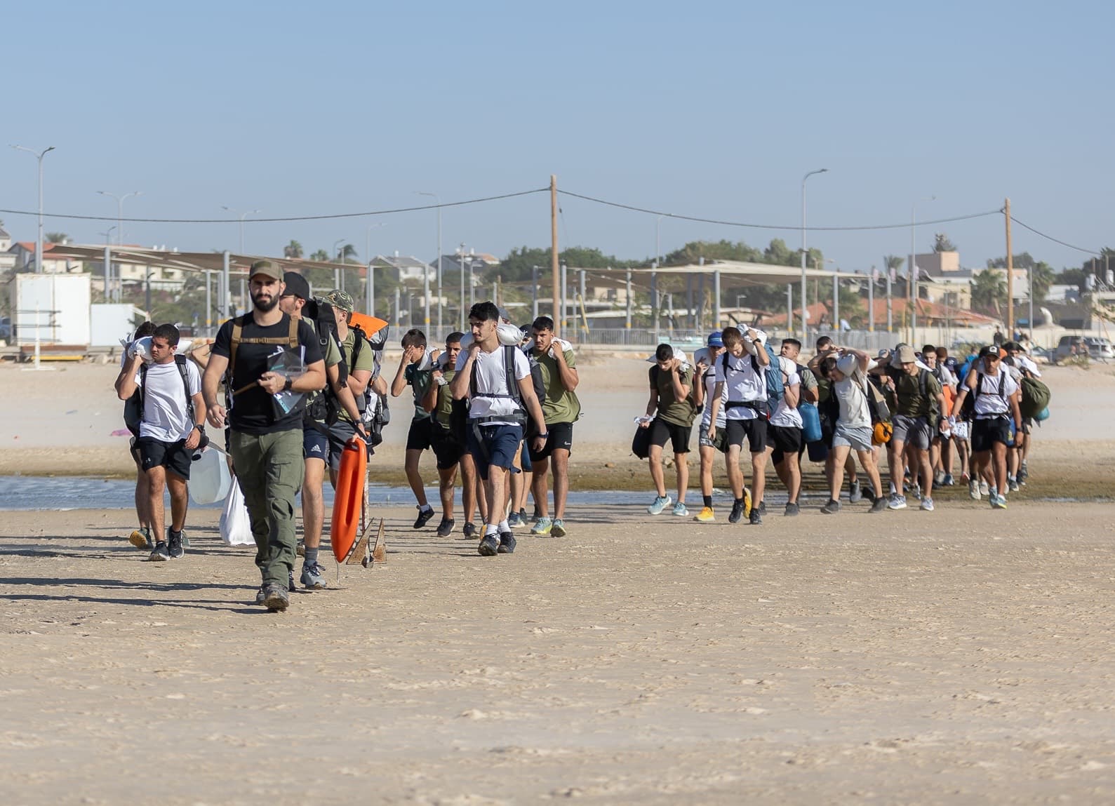 קבוצת חניכים של אדרנלין כושר קרבי צועדת על החוף עם שקים על הכתפיים, ומדריך בראשה
