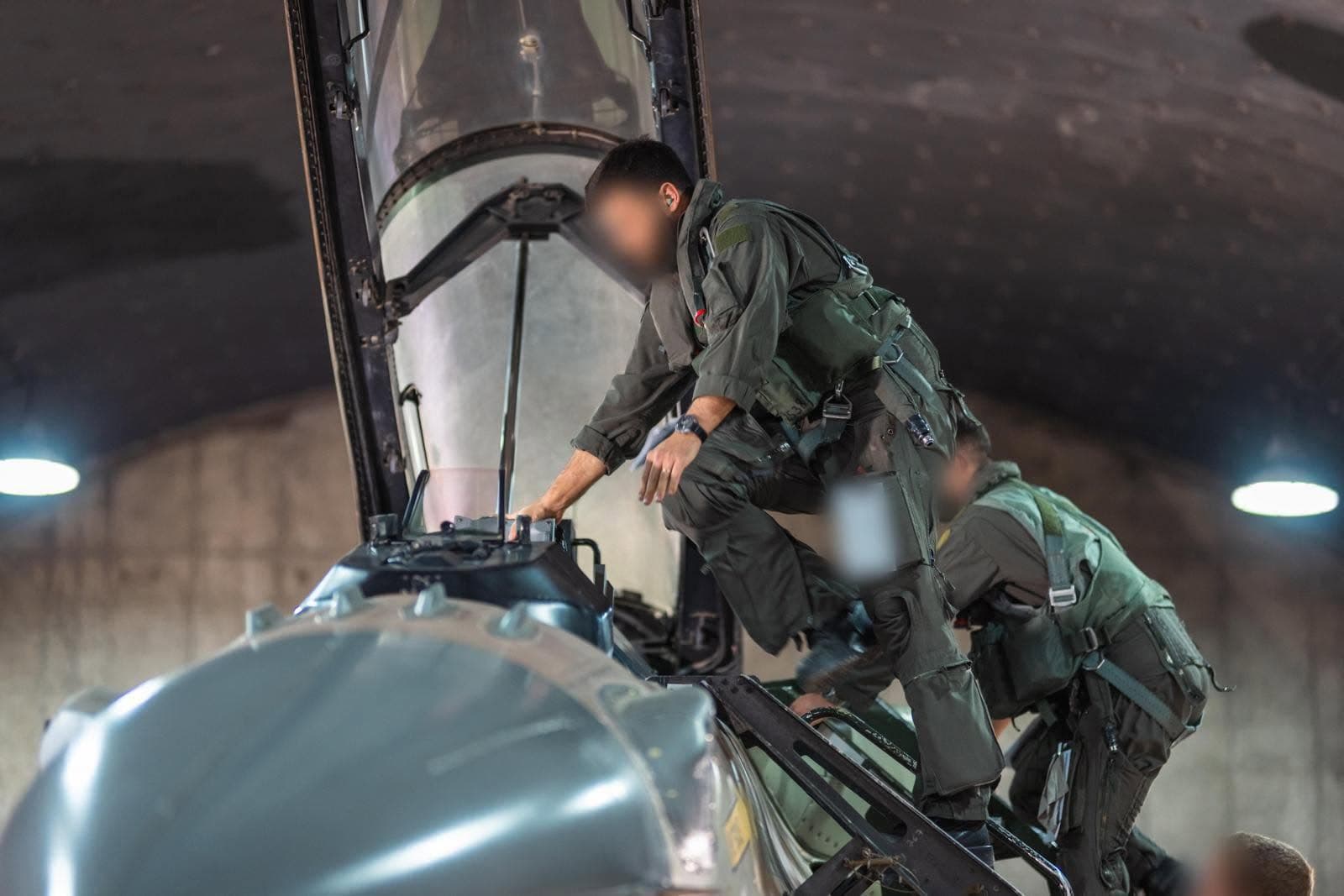 שני אנשי צוות אוויר בסרבלי טיסה עולים בסולם אל תא הטייס של מטוס קרב, בתוך מבנה בטון מקומר