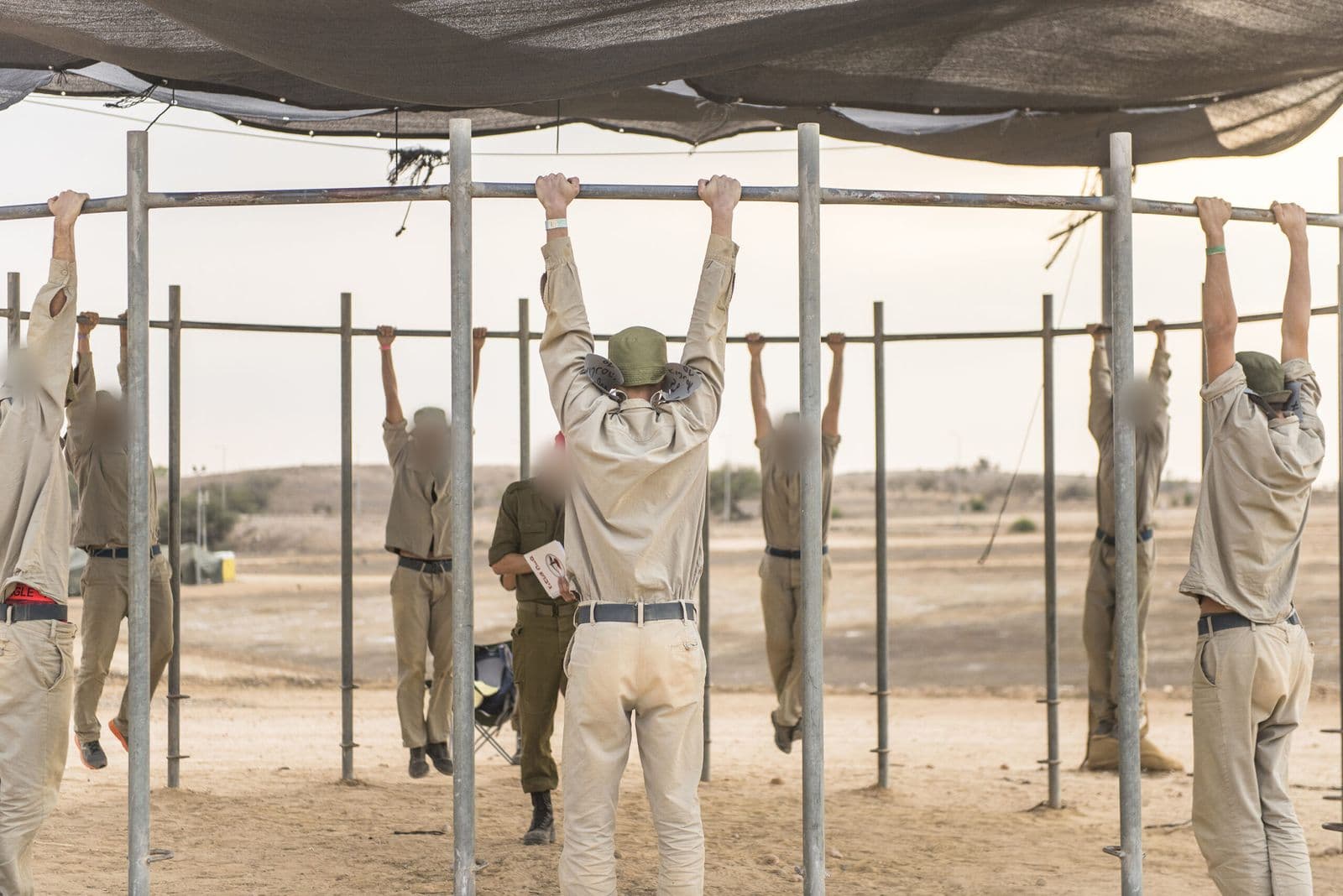 מועמדים בגיבוש טיס תלויים בידיים על מוטות מתכת מתחת לרשת צל, ומגבש עם פנקס עומד ביניהם