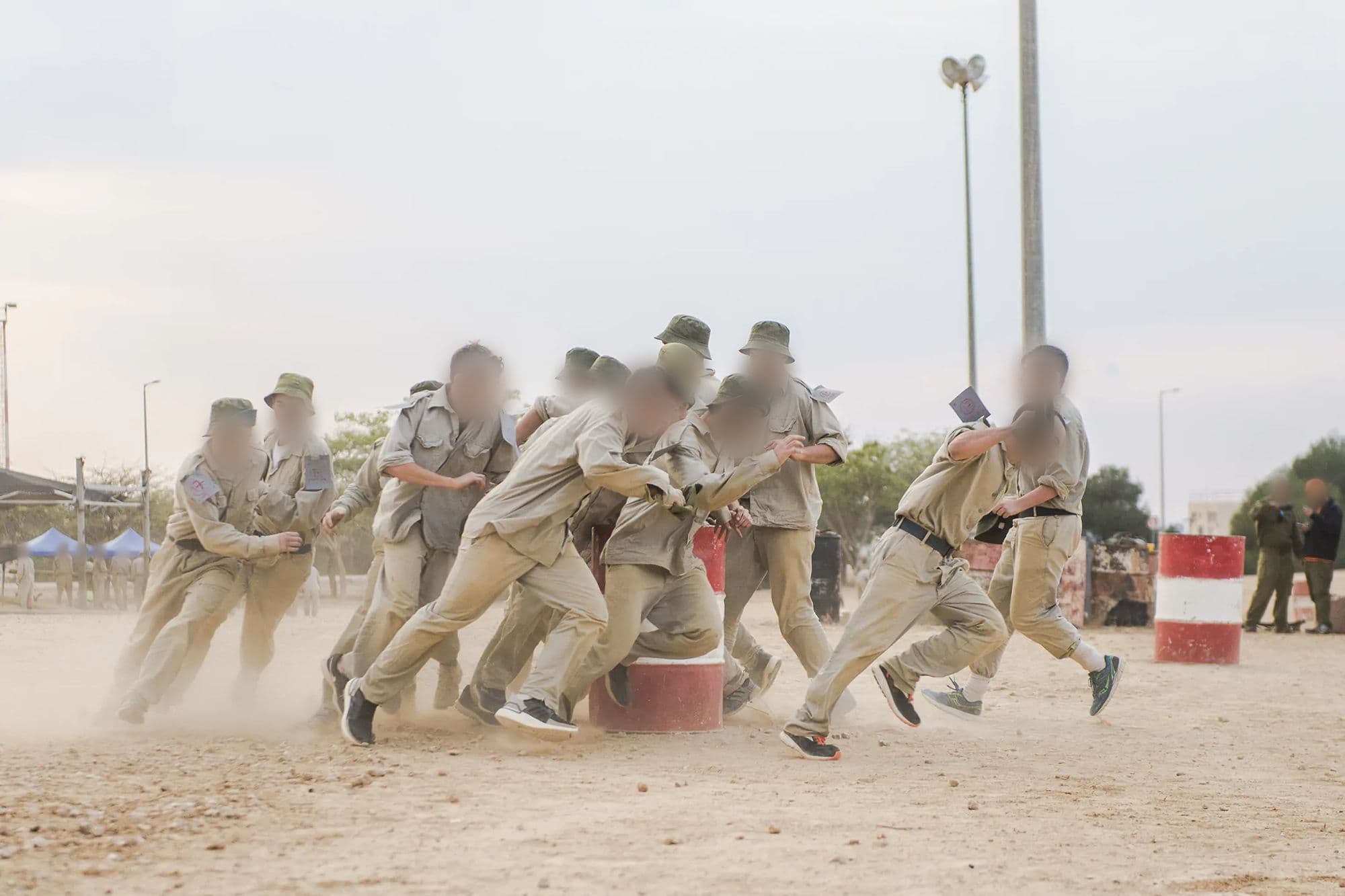 מועמדים בגיבוש טיס, במדים בהירים ובכובעי טמבל, מזנקים בריצה סביב חבית בענן של אבק