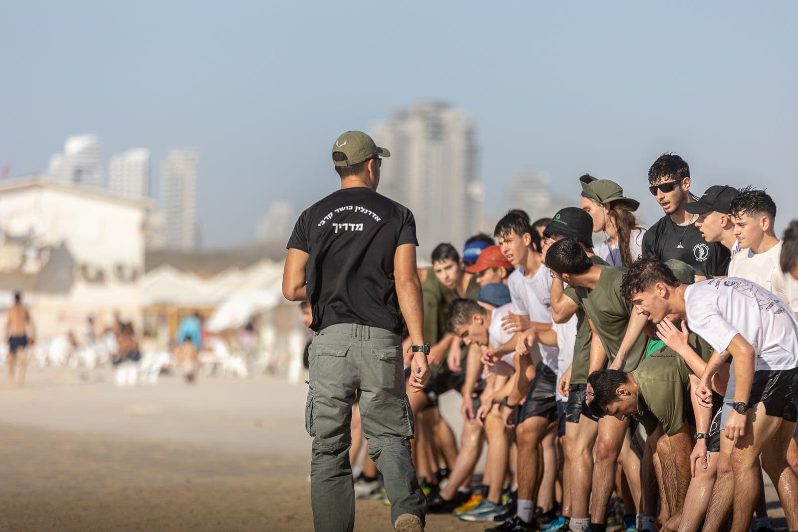 מדריך אדרנלין כושר קרבי עומד מול שורת חניכים על החוף, והחניכים רכונים קדימה וידיהם על הברכיים