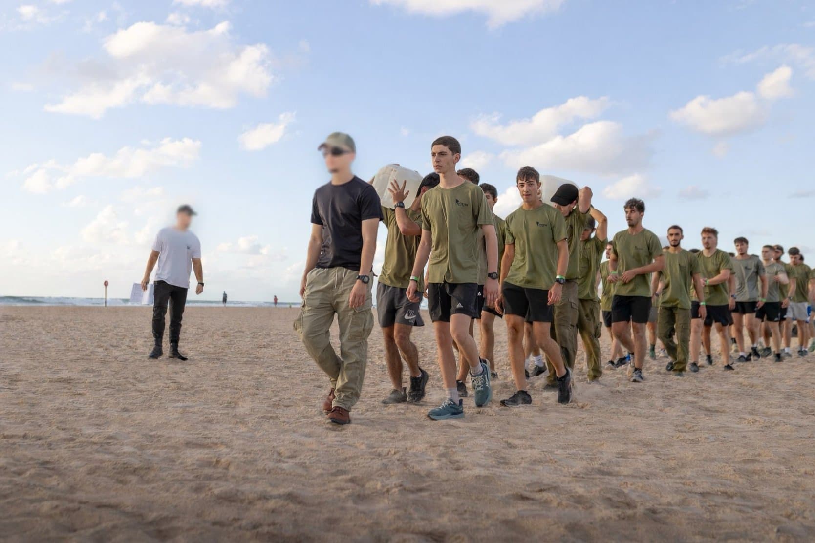 שורה ארוכה של חניכי אדרנלין כושר קרבי בחולצות ירוקות הולכת על חוף הים, חלקם נושאים ג׳ריקנים על הכתפיים, ומדריך הולך בראש השורה