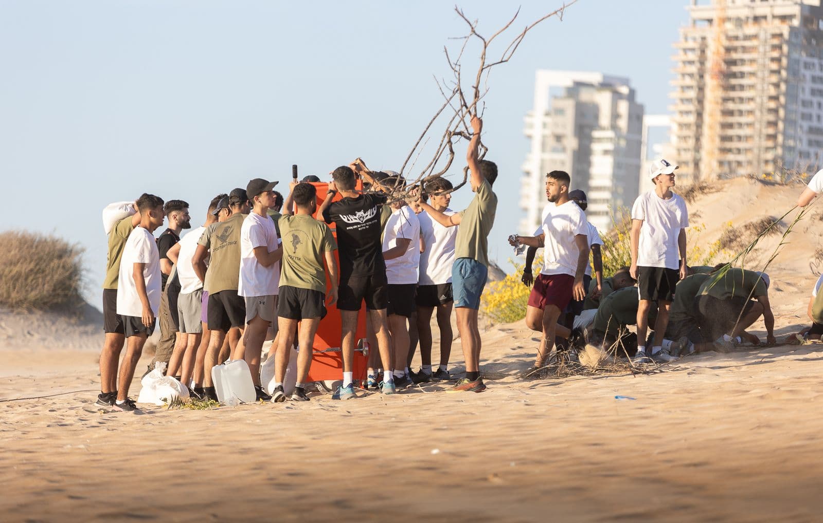 חניכי אדרנלין כושר קרבי עובדים בצוות על החול סביב לוח כתום, ולרגליהם ג׳ריקנים ושקים לבנים
