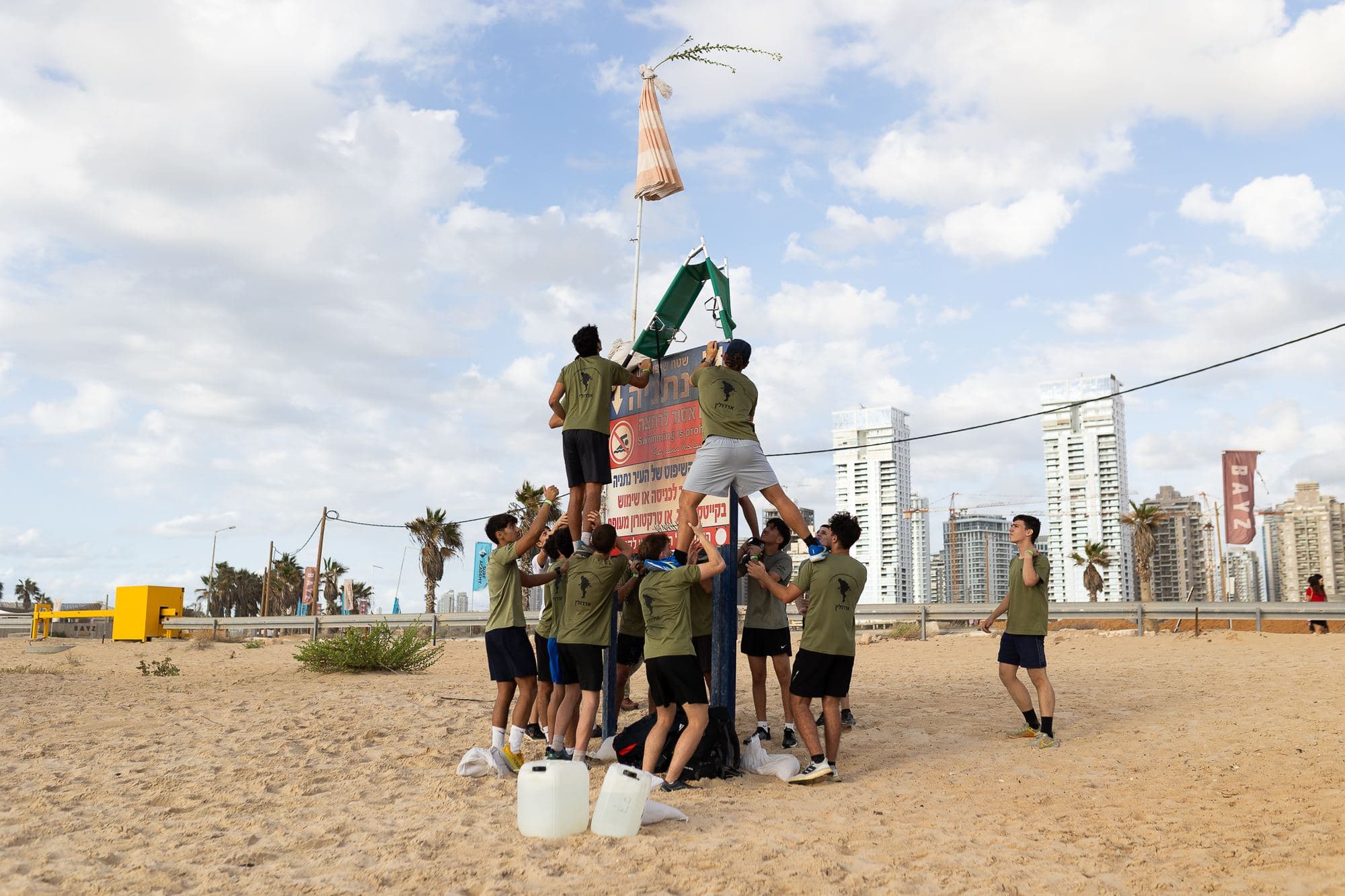 משימת צוות של חניכי אדרנלין כושר קרבי על החוף: שני חניכים עומדים על כתפי חבריהם ומגיעים לאלונקה שבראש שלט